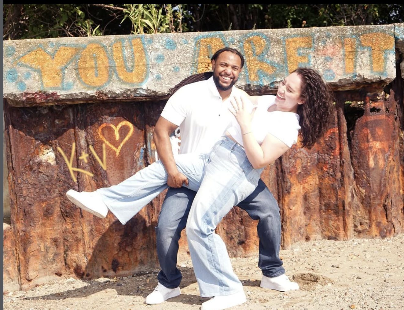 The couple laughing in front of a rusted wall painted with the words YOU ARE IT.