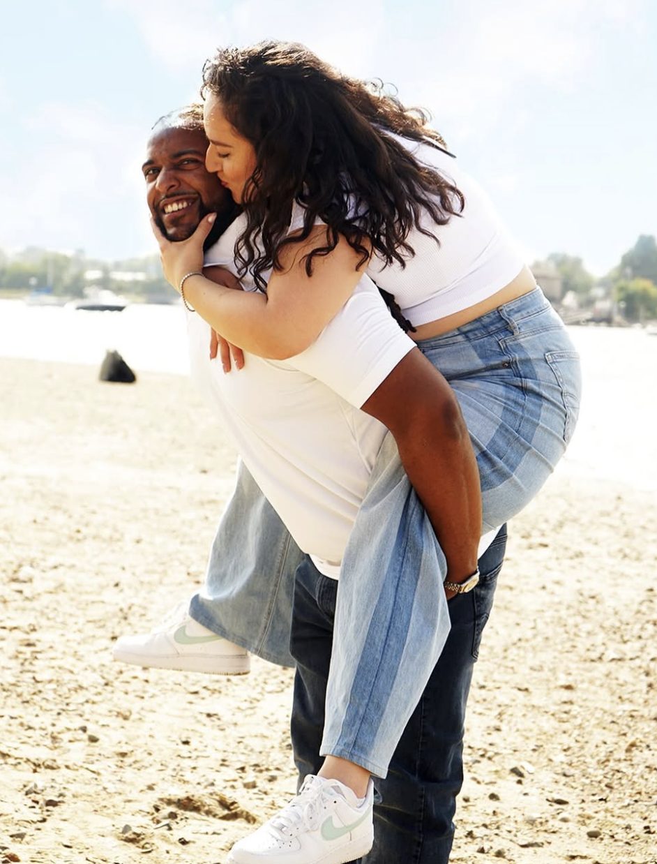 The woman rides piggyback and kisses the man's cheek on a sunny beach.