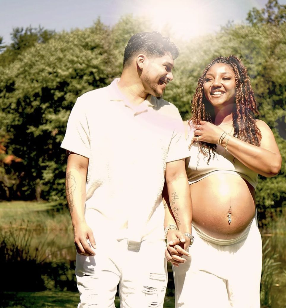 An expecting couple holding hands and smiling at each other in bright sunlight among trees.