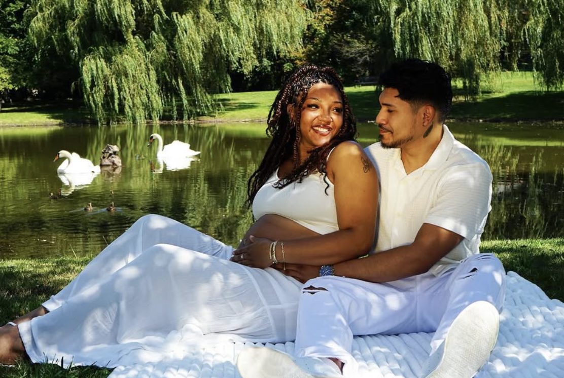 The couple sits on a white blanket by a pond as two swans glide behind them.