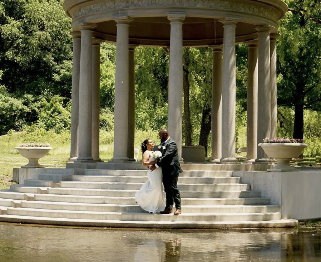 Bride and groom on the steps of a garden temple beside a pond.