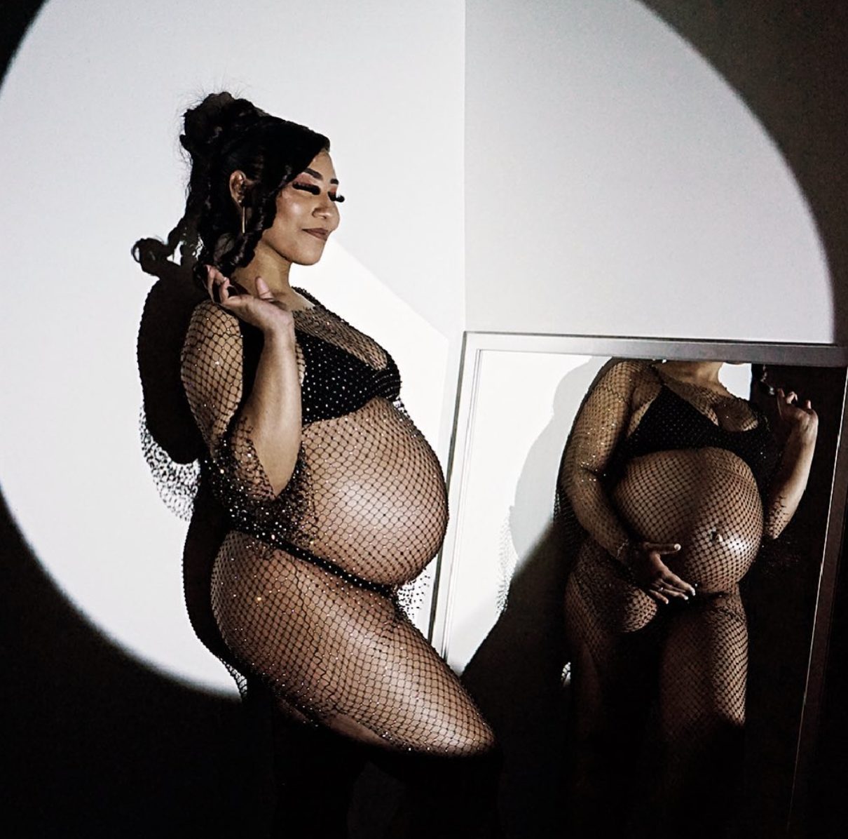 She smiles with eyes closed beside a mirror that reflects her belly.