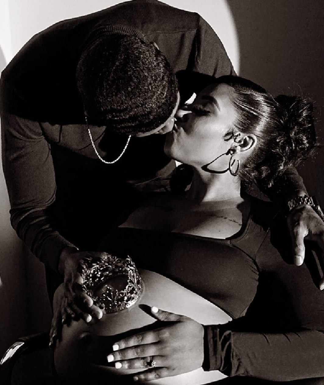 Black and white: her partner leans down to kiss her while a small crown rests on her belly.