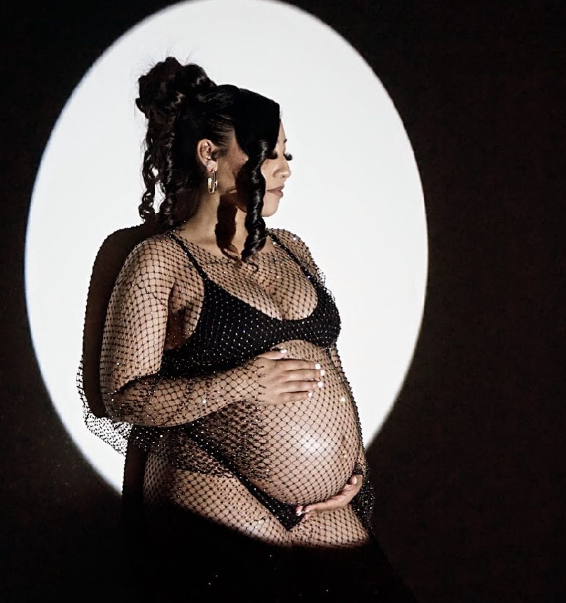 A mother-to-be in a sparkling net dress inside a spotlight.