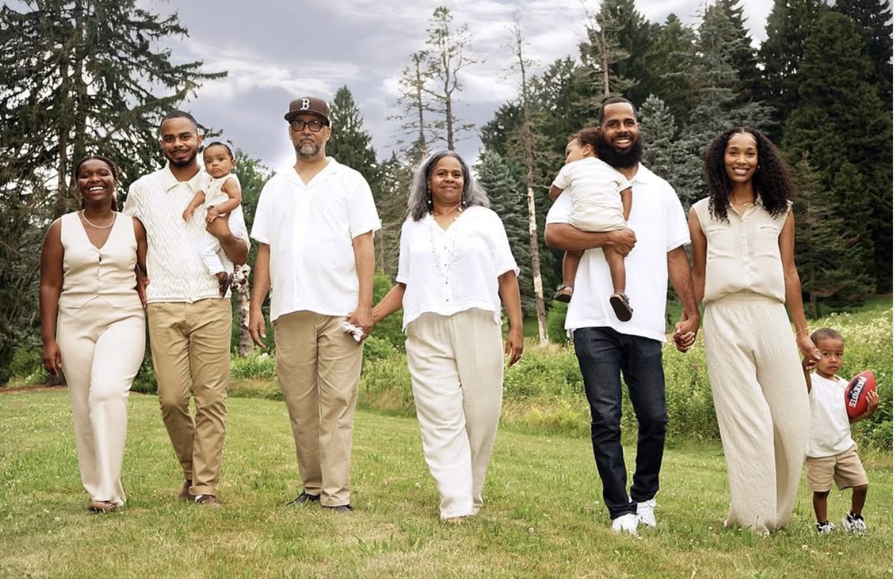 A family of three generations dressed in cream and white walks toward the camera across a meadow.