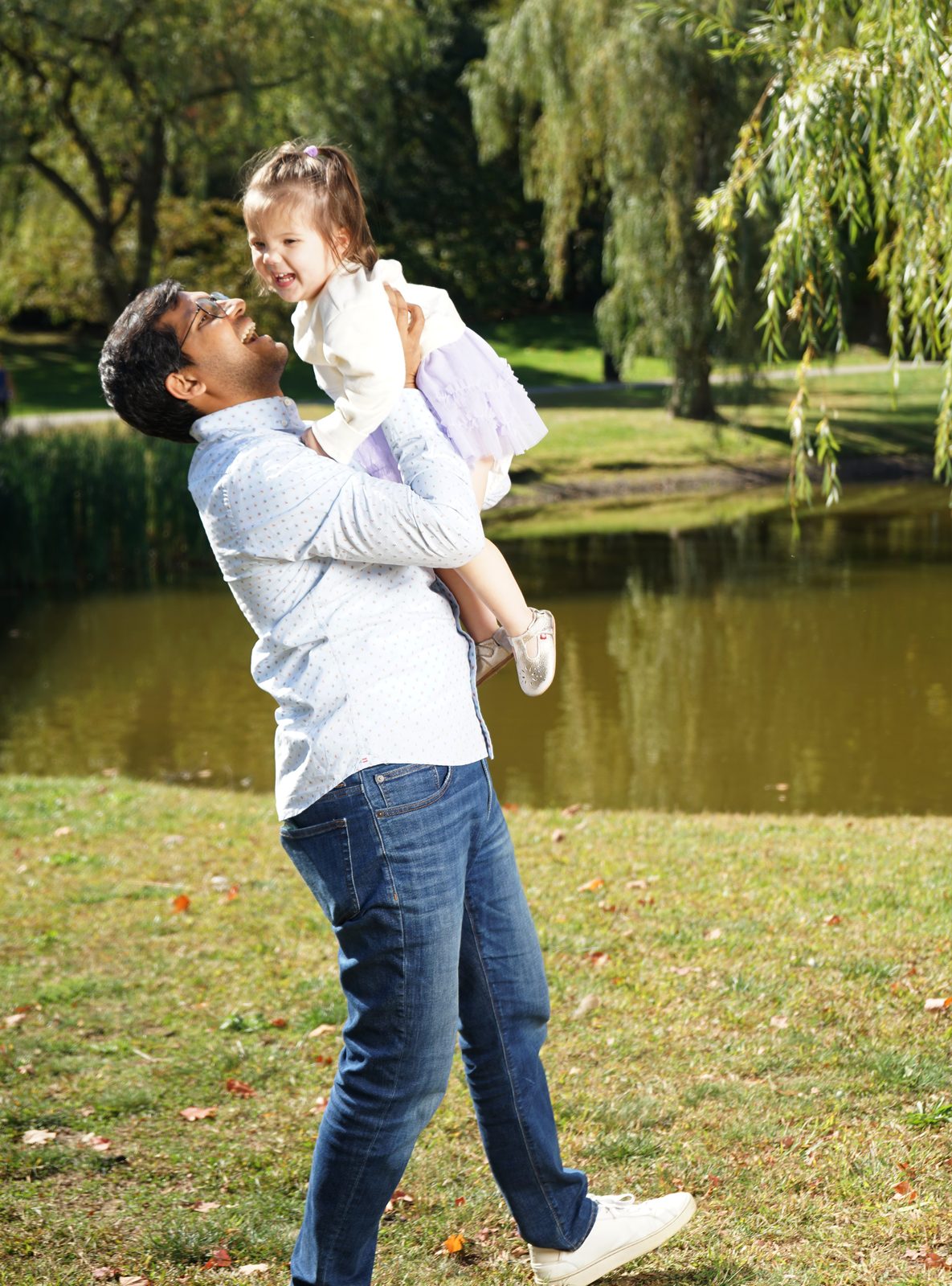 A father lifts his laughing daughter high in the air beside a pond under willow trees.