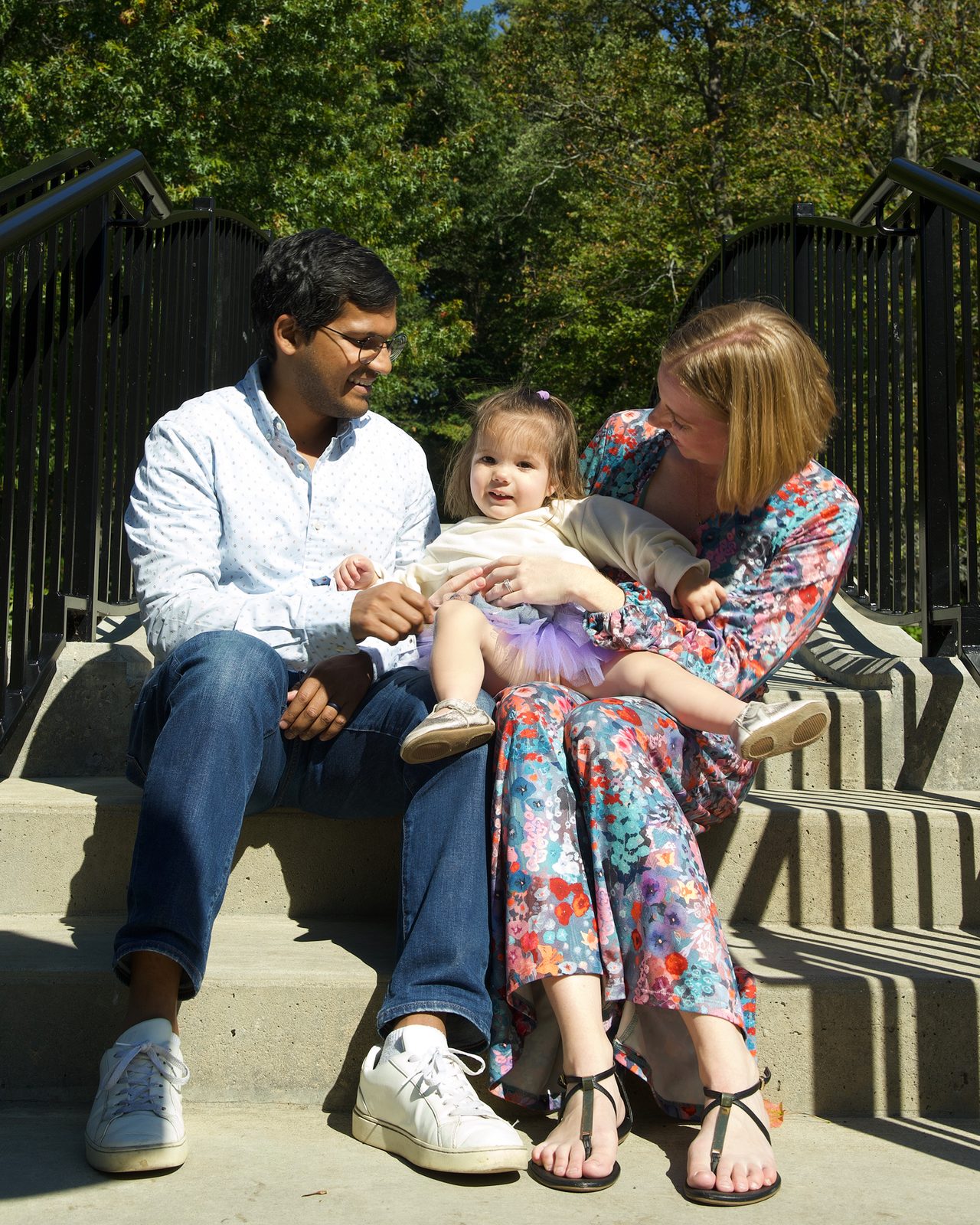 Mother and father sit on sunlit stone steps with their toddler on her lap.