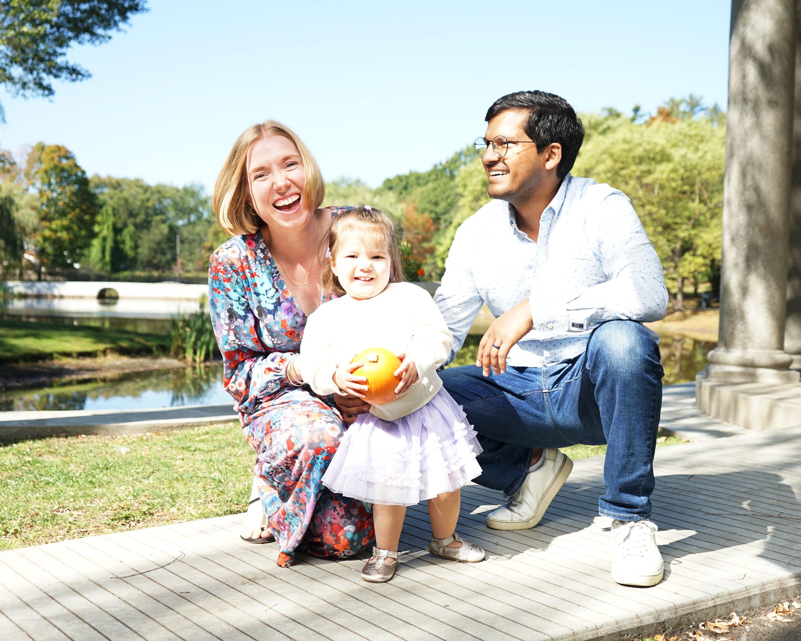 A laughing mother and father crouch beside their toddler, who holds a small pumpkin, by a pond in early autumn.