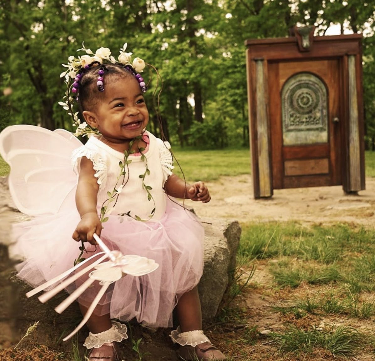 A laughing toddler in fairy wings beside an antique wooden door in the woods.