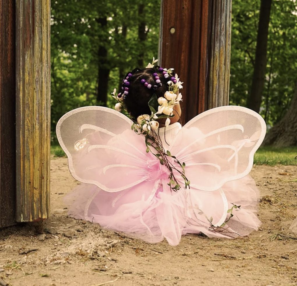 Seen from behind, the little fairy sits in the doorway with her wings spread and vines trailing from her crown.