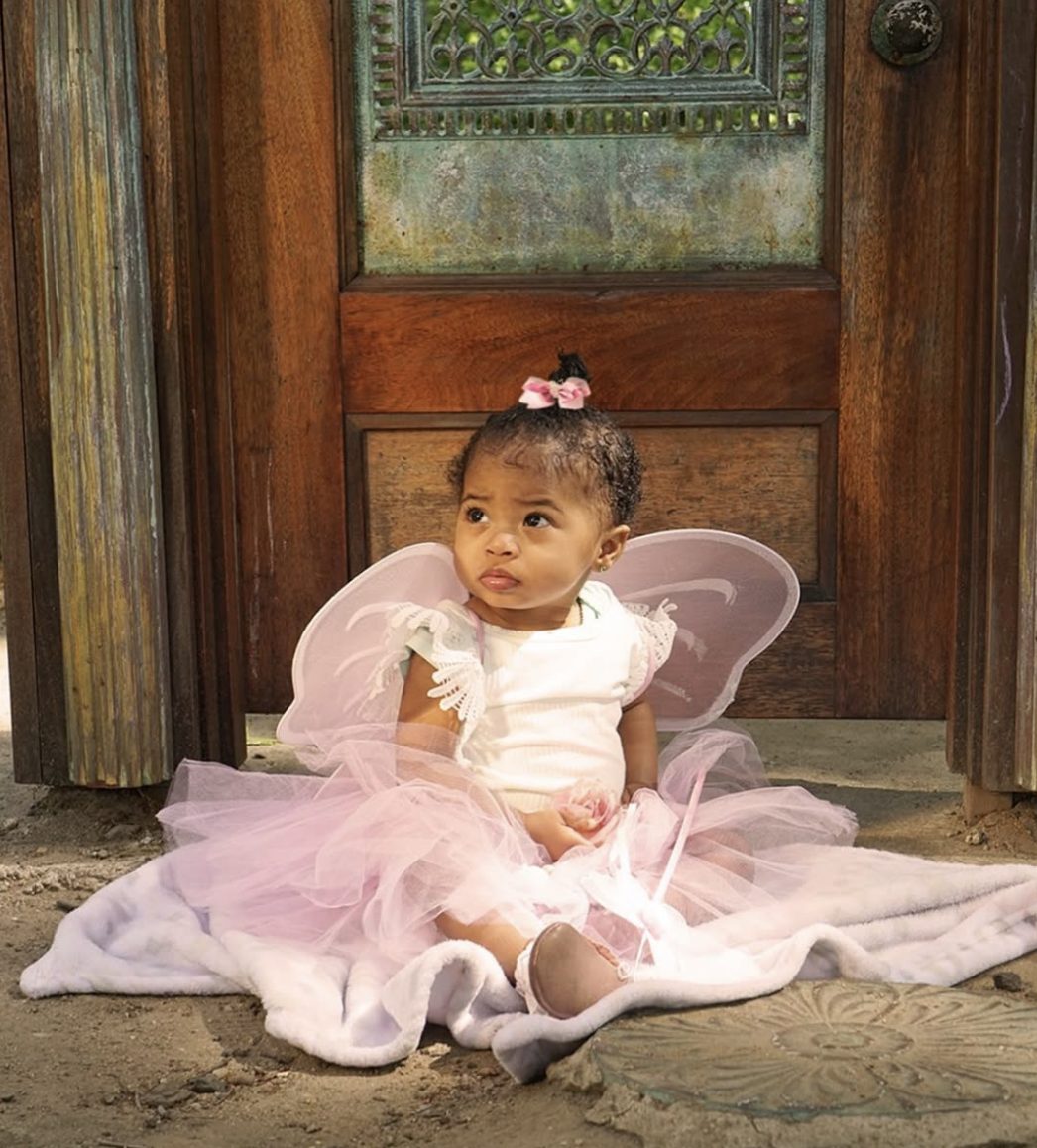 The toddler sits on a white blanket in front of the carved door, looking up thoughtfully.