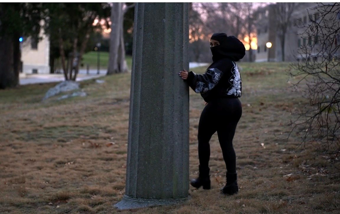 Masked figure in the Edition 01 hoodie beside a stone column at sunset.